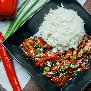A classic Asian dish – rice with teriyaki fried pork wok in a black plate on a wooden background. Close up